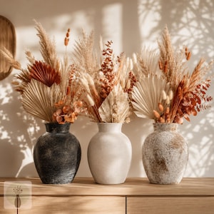 May include: Three rounded vases in black, off-white, and distressed beige, each filled with a dried floral arrangement featuring pampas grass, fan-shaped palm leaves, and rust-colored foliage. The vases have a textured finish and wide bodies.