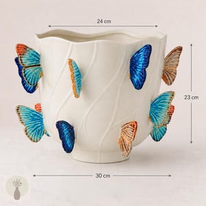 May include: A white ceramic planter featuring raised, three-dimensional butterfly decorations in shades of blue, orange, and brown. The planter has a wavy rim and measures 24 cm in width at the top, 30 cm in width at the base, and 23 cm in height.