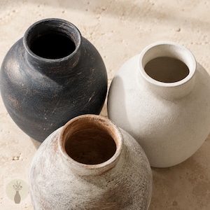 May include: Three round vases with narrow necks and wide bodies. One vase is matte black with visible brushstrokes, one is off-white with a textured finish, and one is a weathered brown with a distressed, light-colored wash over the surface.