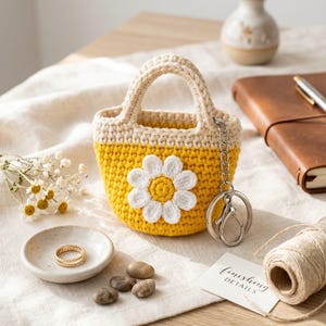 May include: A handmade yellow and cream crochet bag with a white daisy applique. The bag has a small handle and a silver keychain. A small ceramic dish with a gold ring, pebbles, and a tag that says "FINISHING DETAILS" are also present.