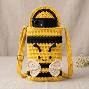 May include: A yellow and black crocheted crossbody bag shaped like a bee, featuring a flap top, two small off-white crocheted wings, embroidered antennae, and a smiling face with black bead eyes. A black smartphone is tucked inside the bag.