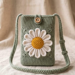Op de afbeelding: Een saliegroene gehaakte schoudertas met een grote witte margriet met een geel hart op de voorzijde. De tas heeft een lange bijpassende schouderband en een enkele houten knoop aan de bovenzijde om de opening van het rechthoekige tasje te sluiten.