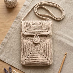 May include: A beige crochet crossbody bag featuring a rectangular body with a geometric pattern, a fold-over flap, and a central circular button closure. The bag includes a long matching crochet strap and is crafted from light tan yarn.