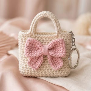 May include: A cream-colored crocheted mini handbag keychain featuring a prominent light pink crocheted bow on the front. The small bag has a structured handle and is attached to a silver-toned metal split ring and chain for secure attachment.
