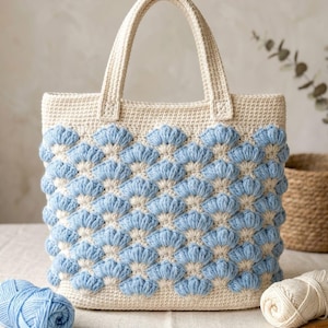 May include: A cream-colored crochet tote bag featuring a textured shell pattern in light blue and cream. The bag has two sturdy top handles and a rectangular structure, showcasing a repeating scalloped design across the front surface.
