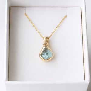 Può includere: Una collana in oro con un ciondolo a goccia in pietra preziosa azzurra traslucida, incastonato in una montatura in oro liscio. Il ciondolo pende da una sottile catenina d'oro, presentato in una scatola quadrata bianca con un inserto bianco.