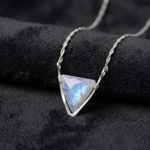 May include: A silver necklace featuring a triangular faceted moonstone pendant with a blue iridescent sheen. The stone is held within a simple silver wire frame that connects to a delicate silver chain on both sides of the triangle.