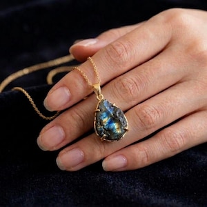 May include: A gold necklace featuring a teardrop-shaped labradorite pendant with a raw, textured surface and iridescent blue and gold flashes. The pendant is held by a delicate gold chain, showcasing the natural stone's unique colour variations.