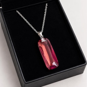 May include: A rectangular, faceted, deep pink crystal pendant suspended from a thin silver-toned metal chain. The pendant features a clear, metallic bail at the top, connecting the crystal to the chain, presented inside a black jewelry box.