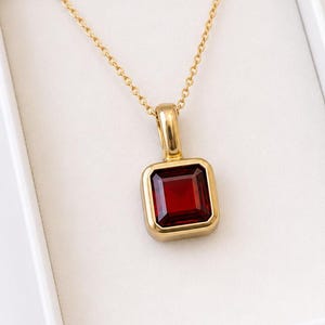 Op de afbeelding: Een gouden hanger aan een ketting met een rechthoekige, dieprode edelsteen gezet in een gladde, gepolijste gouden rand. De hanger hangt aan een fijne gouden ketting en heeft een strak, geometrisch ontwerp met een gefacetteerde steen in het midden.