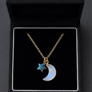 Peut inclure: Un collier à chaîne dorée avec deux pendentifs : une petite étoile à cinq branches avec une finition bleue irisée et un croissant de lune à la surface translucide, blanc laiteux et bleu irisé, présenté dans une boîte noire.