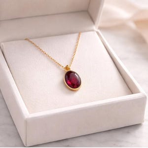 Può includere: Una collana d'oro con un grande pendente ovale in pietra preziosa rosso intenso. La collana è esposta in un portagioie bianco aperto con un interno morbido e strutturato. La catena è delicata.