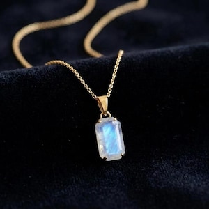 May include: A gold chain necklace featuring a rectangular, faceted moonstone pendant. The gemstone displays a translucent, milky white appearance with a distinct blue adularescence glow, secured by four small gold prongs in a simple setting.