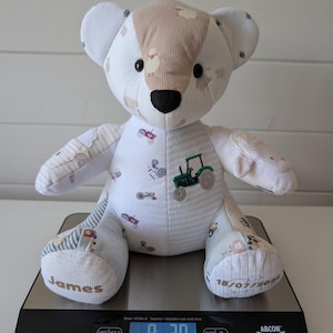 May include: A stuffed teddy bear made from white and beige fabric with a tractor print, featuring a black nose and dark eyes. The name James is embroidered on the left foot and 18/07/2025 on the right foot. It sits on a scale showing 232.5 grams.