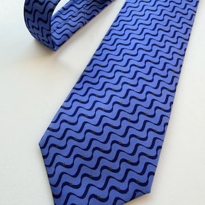 May include: A light blue necktie with a repeating wavy pattern in a darker shade of blue. Small gold-colored dots are interspersed within the design. The tie is made of a smooth fabric and is displayed on a white surface.