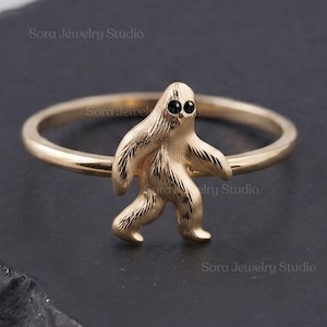 May include: A gold ring featuring a textured, upright creature figure with two small, round black eyes. The figure is positioned in the center of the thin, polished gold band, which forms a complete circle. The design is simple and metallic.