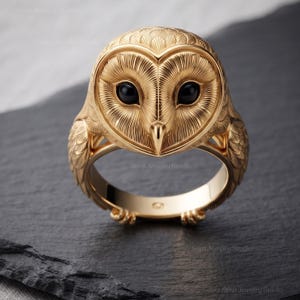 Könnte beinhalten: Ein goldener Ring in Form eines Schleiereulenkopfes mit detaillierten Federgravuren und zwei glatten, runden schwarzen Steinen als Augen. Die Ringschiene zeigt an den Seiten ausgearbeitete Flügelmuster und kleine krallenartige Akzente.