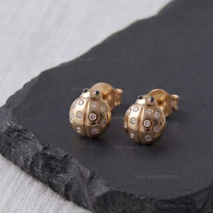 May include: A pair of gold stud earrings shaped like ladybirds, featuring a polished gold finish and small, round, clear stones embedded across the surface of the wings. Each earring has a small, dark, round accent representing the head.