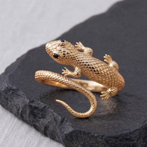 May include: A gold ring shaped like a lizard with a textured, scaled surface. The lizard's body wraps around the finger, and its head features two small, dark blue gemstone eyes. The metal has a polished finish, highlighting the detailed scales.
