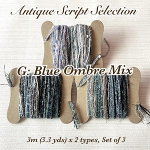 Puede incluir: Tres carretes de hilo azul ombre mix en una exhibición de cartón. El hilo es una mezcla de azules, grises y blanco. El texto "Antique Script Selection" y "G: Blue Ombre Mix" son visibles. El hilo es 3m x 2 tipos, juego de 3.