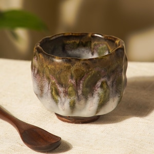 May include: A handmade ceramic tea bowl with a distinctive, textured finish. The bowl displays a mix of earthy colours: brown, green, and white, with a hint of red. A small, dark wooden spoon is placed close by. Ideal for tea lovers.