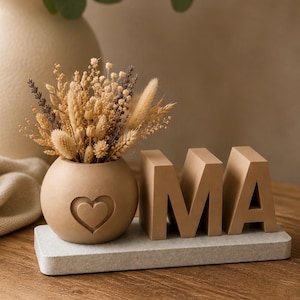 May include: A decorative arrangement featuring a small, round vase with a heart cutout, filled with dried flowers. The word "OMA" is displayed in block letters on a rectangular base. The color palette is neutral, with beige and brown tones.