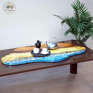 May include: A unique, handcrafted wooden coffee table with a resin inlay. The table features a blue, yellow, and brown design, resembling a beach scene. A black tray with two white teacups and a teapot are on the table. A black teapot and a silver teapot are also on the table.