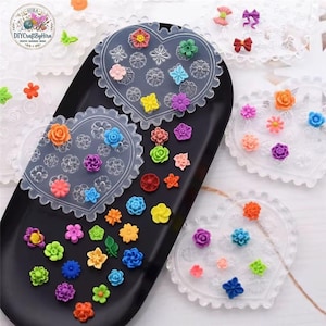 May include: A collection of colorful, miniature flower-shaped embellishments in various sizes and hues, including orange, yellow, blue, and purple. The flowers are arranged on a black tray and heart-shaped molds, perfect for crafting projects.