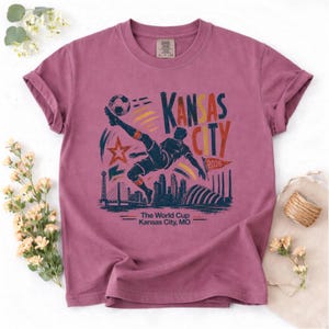 May include: A mauve-colored t-shirt featuring a soccer player kicking a ball, with the text "Kansas City 2026" and a cityscape silhouette. The shirt also includes the text "The World Cup Kansas City, MO."