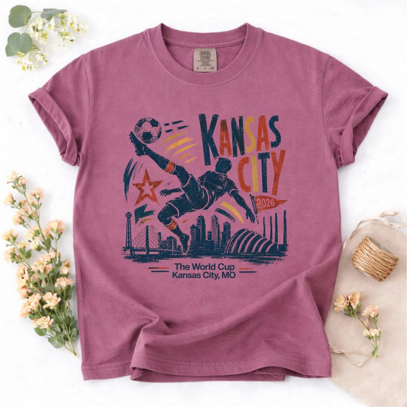 May include: A mauve-colored t-shirt featuring a soccer player kicking a ball, with the text "Kansas City 2026" and a cityscape silhouette. The shirt also includes the text "The World Cup Kansas City, MO."