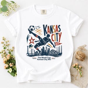 May include: White t-shirt featuring a graphic of a soccer player kicking a ball, with the words "Kansas City 2026" in red, blue, and orange. The shirt also includes the text "The World Cup Kansas City, MO".