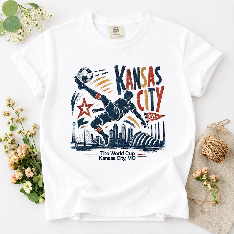 May include: White t-shirt featuring a graphic of a soccer player kicking a ball, with the words "Kansas City 2026" in red, blue, and orange. The shirt also includes the text "The World Cup Kansas City, MO".