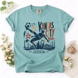May include: A light blue t-shirt featuring a graphic of a soccer player kicking a ball, with the text "Kansas City 2026". The design includes a star, skyline, and the words "The World Cup Kansas City, MO".