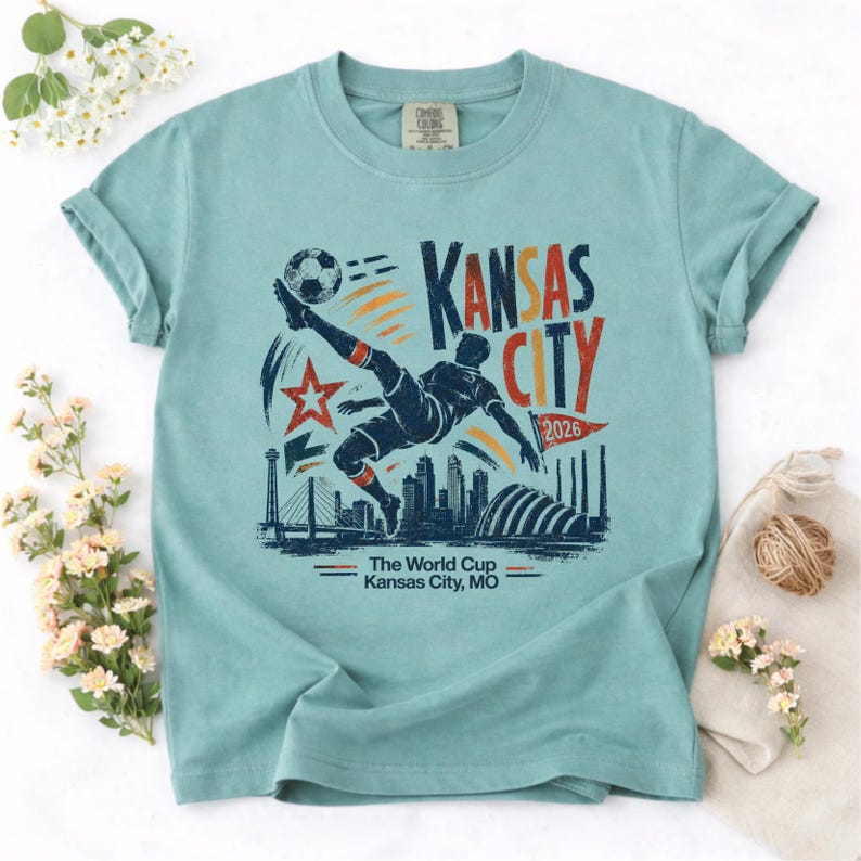 May include: A light blue t-shirt featuring a graphic of a soccer player kicking a ball, with the text "Kansas City 2026". The design includes a star, skyline, and the words "The World Cup Kansas City, MO".