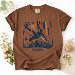 May include: Brown t-shirt featuring a graphic of a soccer player kicking a ball, with the text "Kansas City" and "The World Cup Kansas City, MO". The design includes a star, skyline, and bridge.