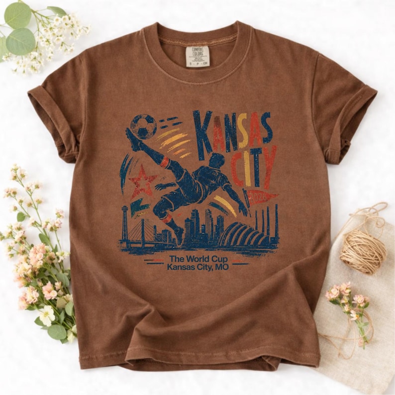 May include: Brown t-shirt featuring a graphic of a soccer player kicking a ball, with the text "Kansas City" and "The World Cup Kansas City, MO". The design includes a star, skyline, and bridge.