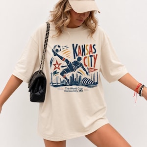 May include: Cream-colored t-shirt featuring a graphic of a soccer player kicking a ball, with the words "Kansas City 2026." The design includes a star, skyline, and the text "The World Cup Kansas City, MO."
