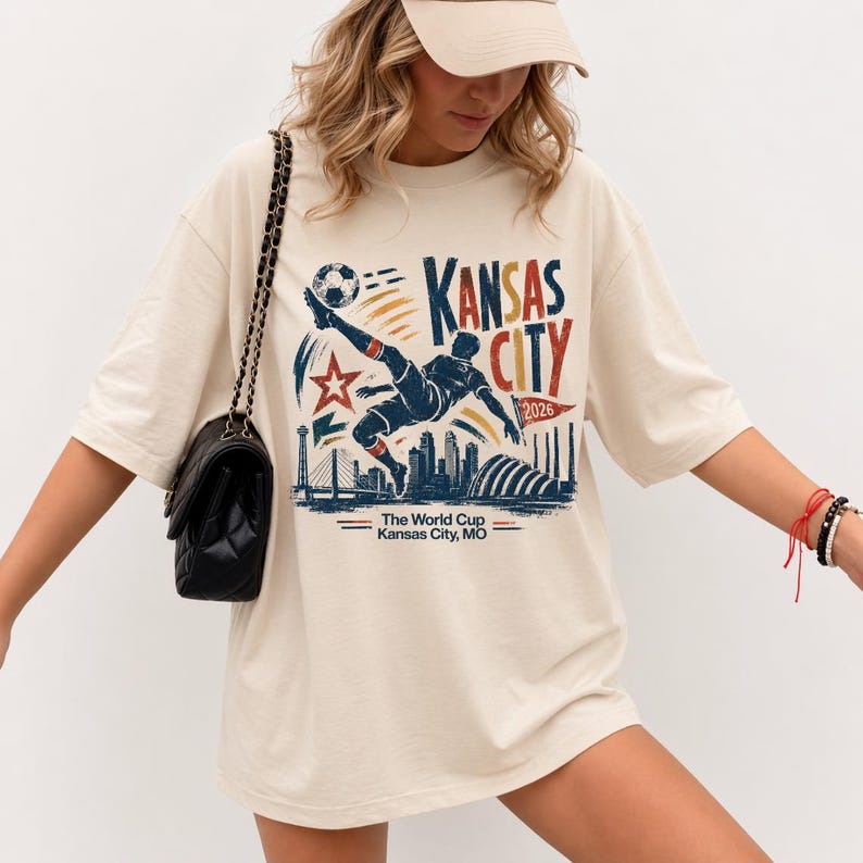 May include: Cream-colored t-shirt featuring a graphic of a soccer player kicking a ball, with the words "Kansas City 2026." The design includes a star, skyline, and the text "The World Cup Kansas City, MO."