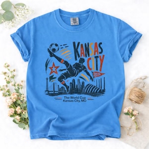 May include: A light blue t-shirt featuring a soccer player kicking a ball, with the text "KANSAS CITY" and "2026". The design includes a star, a bridge, and a cityscape. The text "The World Cup Kansas City, MO" is also present.
