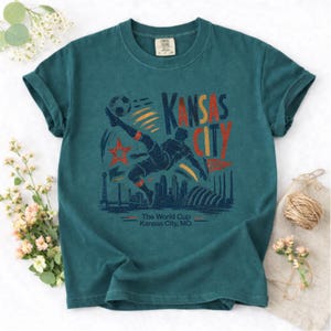 May include: Teal-colored t-shirt featuring a graphic of a soccer player kicking a ball, with the words "Kansas City" in red and orange. The design includes a star, city skyline, and the text "The World Cup Kansas City, MO."