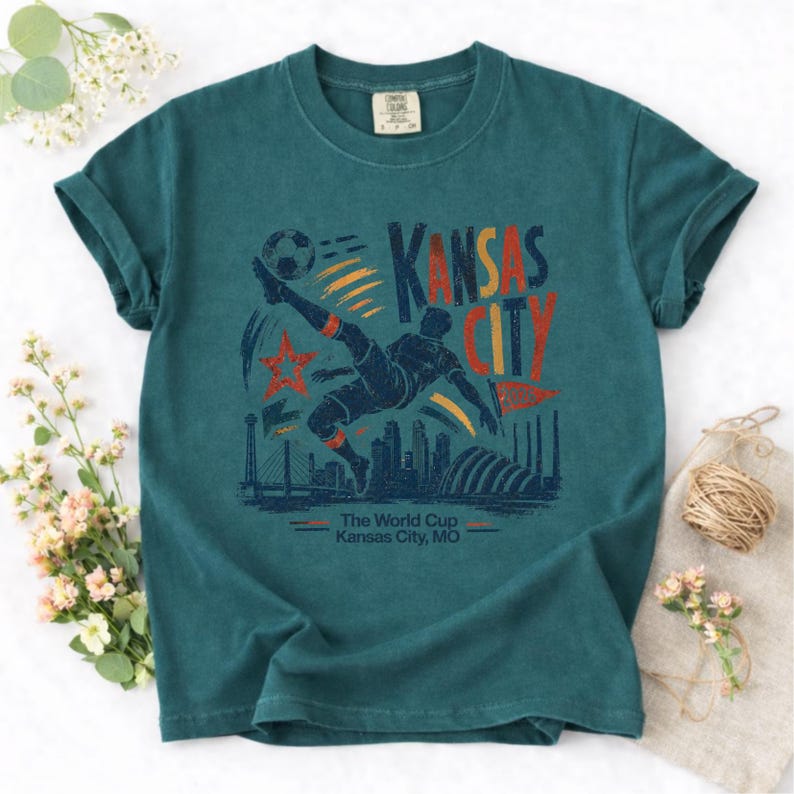 May include: Teal-colored t-shirt featuring a graphic of a soccer player kicking a ball, with the words "Kansas City" in red and orange. The design includes a star, city skyline, and the text "The World Cup Kansas City, MO."