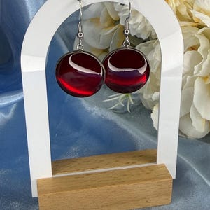 Op de afbeelding: Een paar ronde, rode oorbellen met een glanzende afwerking, hangend aan zilverkleurige haken. De oorbellen worden gepresenteerd op een witte, boogvormige standaard met een houten basis, tegen een achtergrond van witte bloemen en blauwe satijn.
