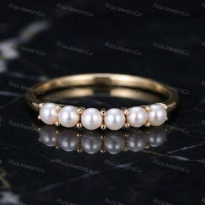 Puede incluir: Un anillo de banda dorada con una hilera de seis pequeñas perlas blancas redondas dispuestas horizontalmente en la parte frontal. El metal tiene un acabado pulido y las perlas están espaciadas uniformemente y aseguradas con pequeñas garras doradas.