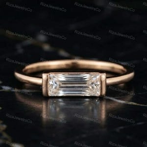 May include: A rose gold ring featuring a single horizontal baguette-cut clear gemstone set in a bezel setting. The band is smooth and circular, showcasing a clean, minimalist design with a polished metallic finish and a central rectangular stone.