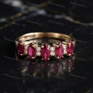May include: A gold band ring featuring five marquise-cut red rubies arranged in a row, interspersed with small round white diamonds. The ring has a polished metal finish and a symmetrical design with stones set in individual prongs along the band.