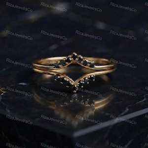 May include: A gold chevron-shaped ring featuring a series of small, round, black stones set along the V-shaped band. The ring has a polished metallic finish and a pointed design that creates a distinct angular silhouette across the finger.