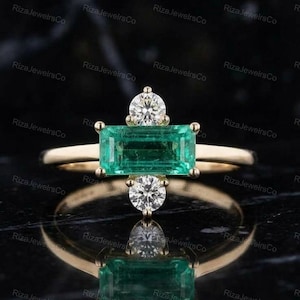 May include: A gold ring featuring a horizontal emerald-cut green gemstone at the center, flanked by a round brilliant-cut white diamond above and below the main stone, set on a polished gold band.