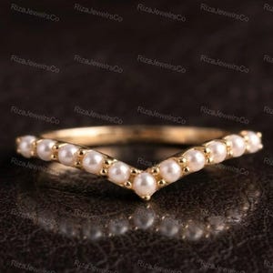 Puede incluir: Un anillo de oro en forma de V con una hilera de pequeñas perlas blancas redondas engastadas a lo largo de la banda. El anillo tiene un acabado metálico pulido y un diseño central en punta, con las perlas espaciadas uniformemente en la curva.