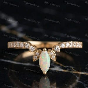 May include: A gold chevron-shaped ring featuring a central marquise-cut opal gemstone flanked by two small leaf-shaped accents, each set with tiny round diamonds. The band is also adorned with a row of small round diamonds along both sides.