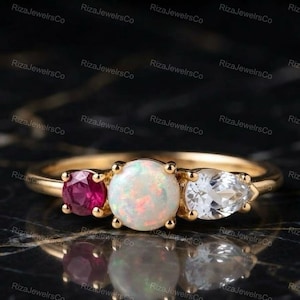 May include: A gold ring featuring a central round cabochon opal, flanked by a round-cut deep pink gemstone on one side and a pear-cut clear white gemstone on the other, all set in a polished gold band with a smooth, rounded profile.